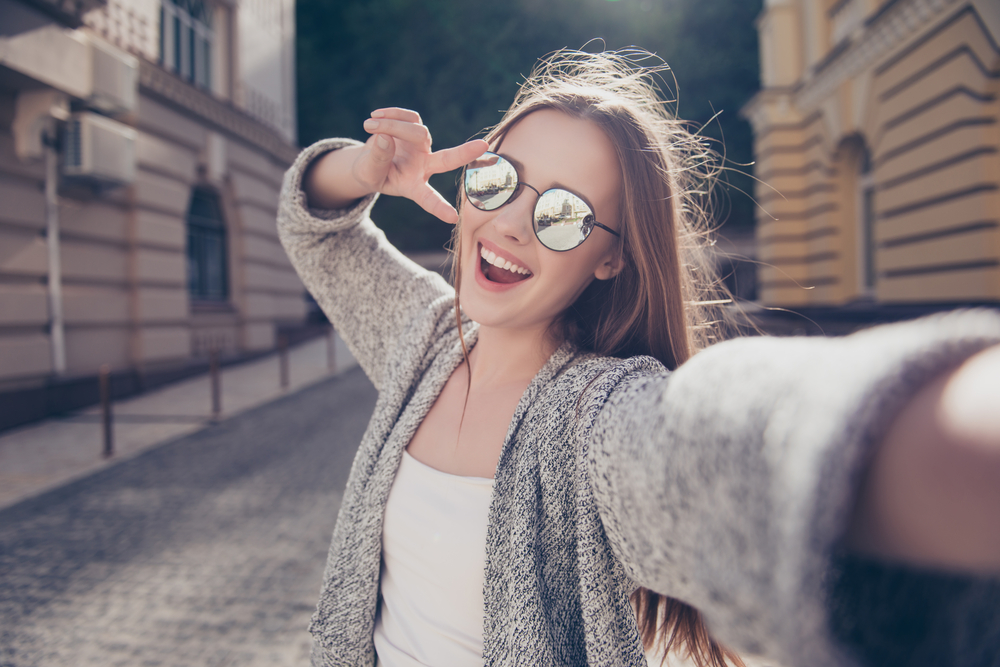 Woman Taking Selfie Wearing Reflective Sunglasses - Desert Vision Eye Care Smiling woman wearing reflective sunglasses while taking a selfie outdoors, representing stylish eyewear and clear vision from Desert Vision Eye Care - Desert Vision Eye Care