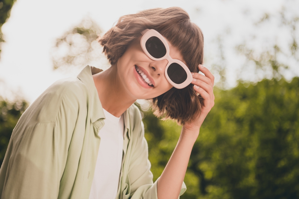 Woman Wearing Trendy Sunglasses Smiling Outdoors - Desert Vision Eye Care Smiling woman wearing stylish sunglasses in a sunny outdoor setting, representing fashionable eyewear and clear vision from Desert Vision Eye Care - Desert Vision Eye Care