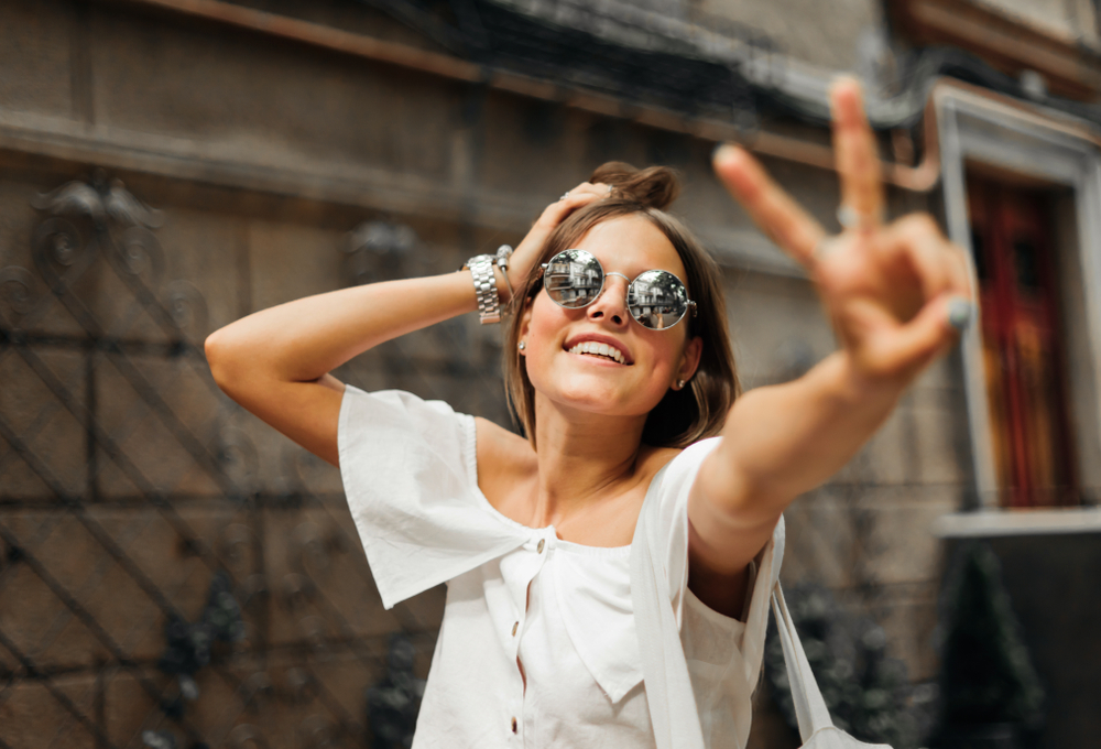 Smiling woman wearing reflective sunglasses and making a peace sign outdoors, representing stylish eyewear and clear vision from Coachella Valley Optometry Indio - Coachella Valley Optometry Indio