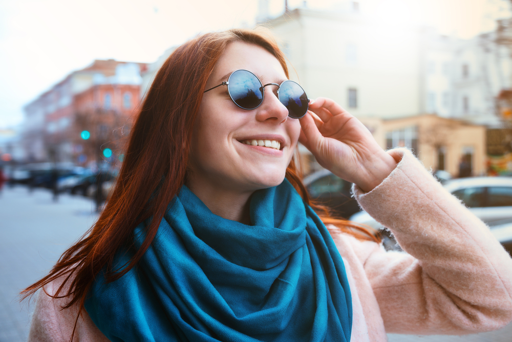Smiling woman adjusting her sunglasses outdoors in an urban setting, representing fashionable eyewear and vision care from an optometrist in Palm Desert - Optometrist Palm Desert