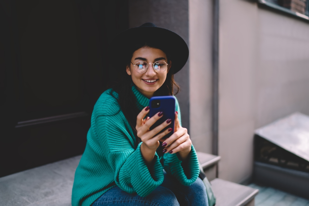 Woman Wearing Eyeglasses Using Smartphone Outdoors - Desert Vision Eye Care Smiling woman wearing eyeglasses and a hat while using her smartphone, representing clear vision and modern eyewear from Desert Vision Eye Care - Desert Vision Eye Care