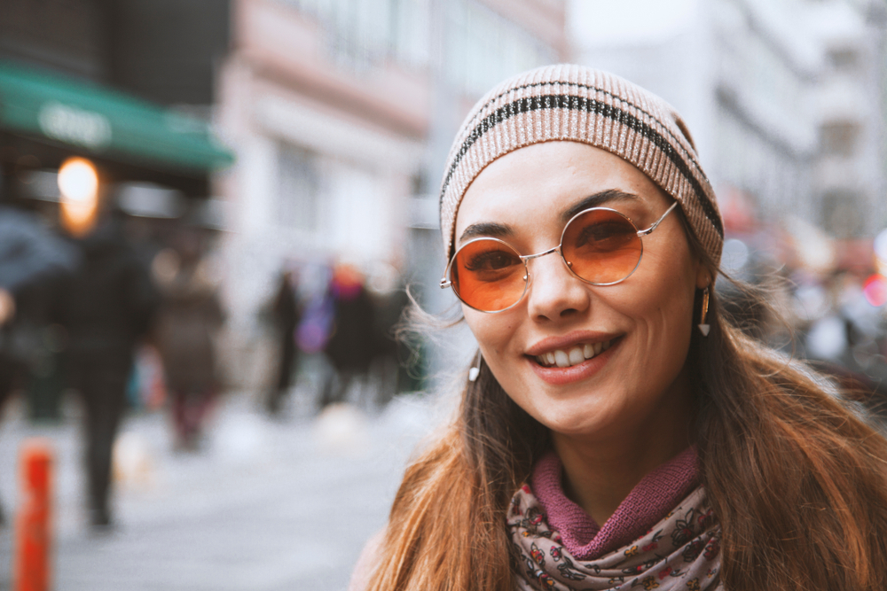 Woman Wearing Fashionable Tinted Glasses in City - Optometrist Indio CA Smiling woman wearing tinted eyeglasses and a beanie in an urban setting, representing stylish vision solutions from an optometrist in Indio CA - Optometrist Indio CA