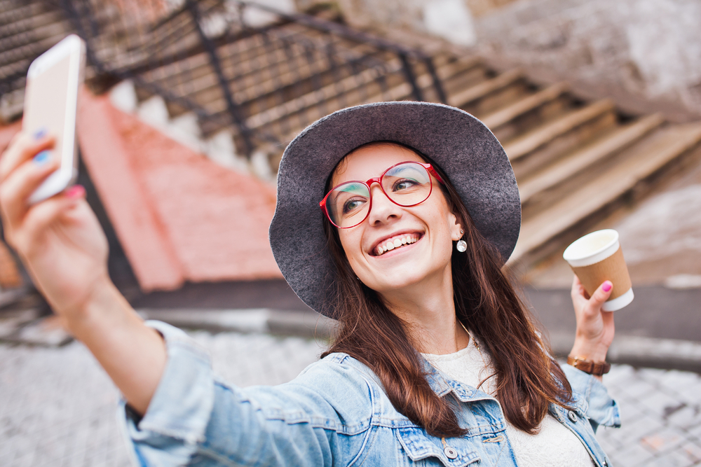 Woman Wearing Stylish Glasses Taking Selfie Outdoors - Optometrist Indio CA Smiling woman wearing trendy eyeglasses and a hat while taking a selfie, representing fashionable eyewear and clear vision from an optometrist in Indio CA - Optometrist Indio CA