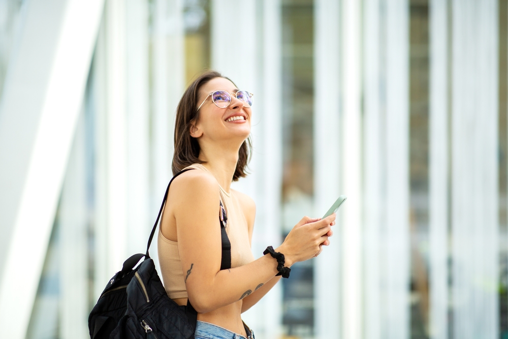 Smiling woman wearing eyeglasses and holding a smartphone outdoors, representing clear vision and stylish eyewear from an optometrist in Palm Desert - Optometrist Palm Desert