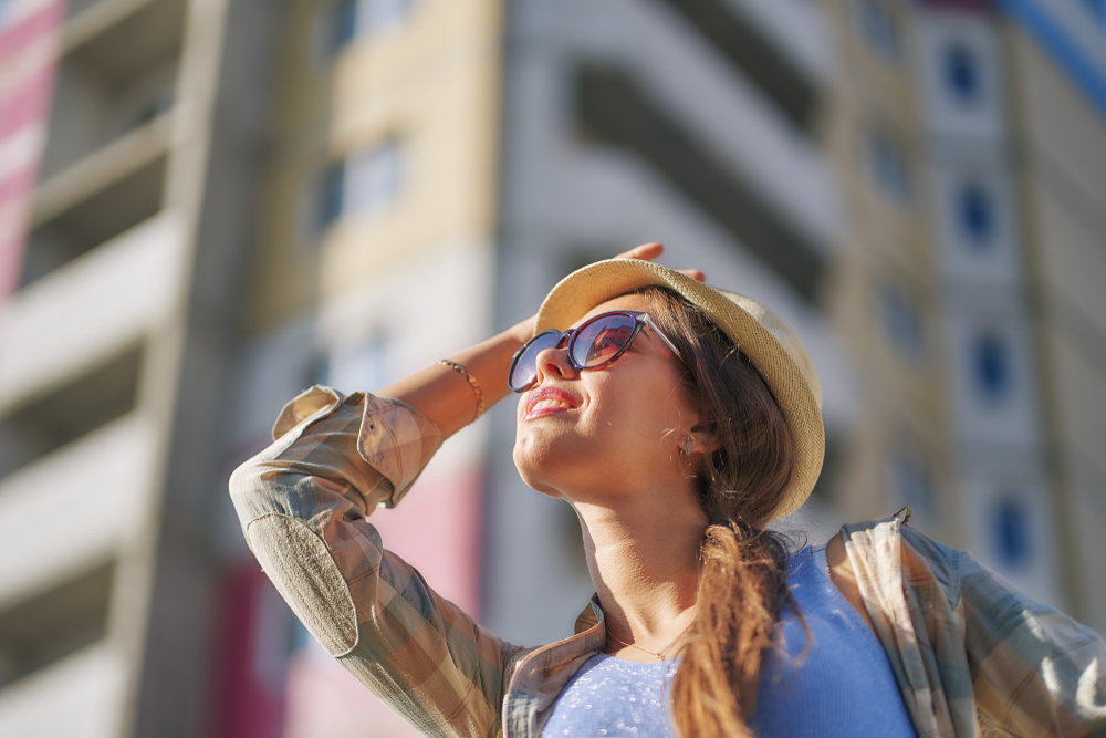 Woman Wearing Sunglasses Enjoying Bright Sunlight Outdoors - Optometrist Indio CA Woman wearing sunglasses and a hat looking up in bright sunlight near buildings, representing UV protection and quality eyewear from an optometrist in Indio CA - Optometrist Indio CA